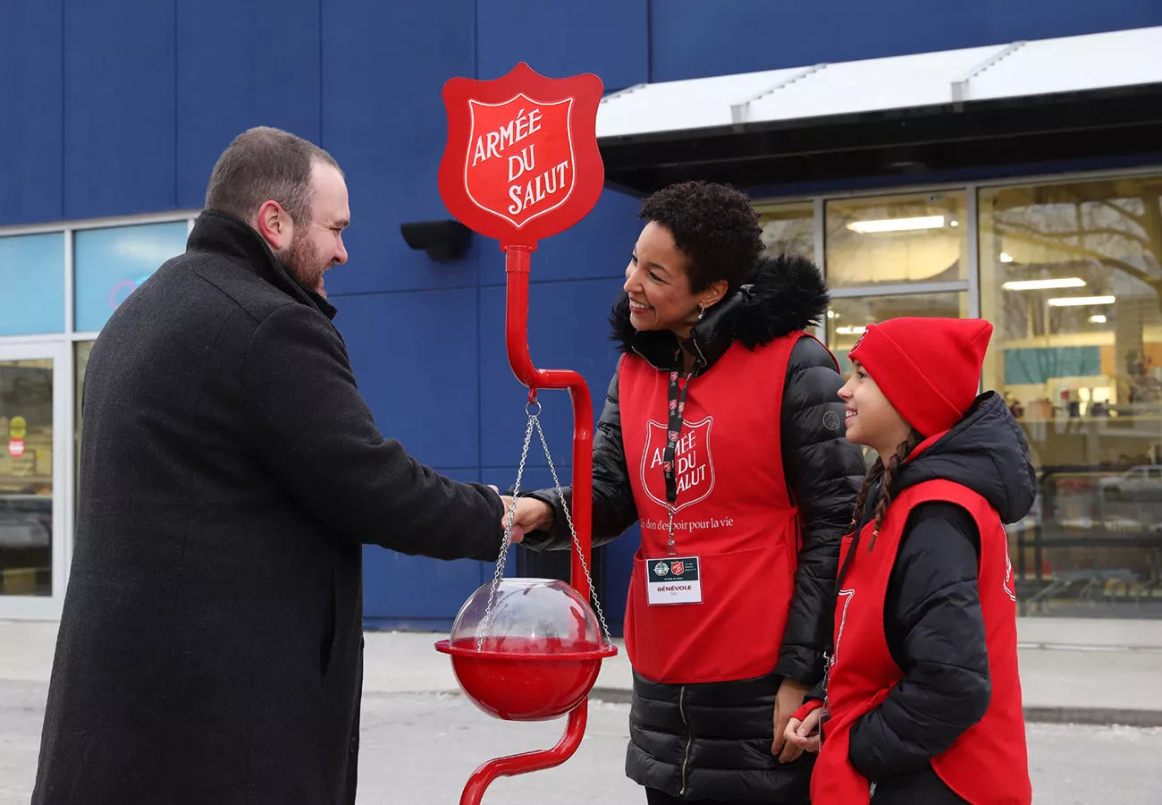 A man donates money to a Salvation Army collection stand outside a building with blue walls and large windows. Two volunteers wearing red vests and hats assist with the donation. The stand features a red sign with the text 'Armée du Salut' and a hanging collection bucket.